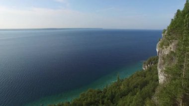 Uçan bir dron altında çam ağaçları ve mavi sular olan bir uçurumdan uçuyor.