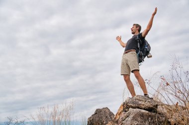 Kafkasyalı yürüyüşçü kayalık dağın zirvesinde duruyor ve mutlu.