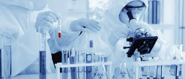 Biyolojik tehlike koruma kıyafet analizi yapan bilim adamı. Laboratuvarda Coronavirus covid-19 kan örneği tüpü tutan başka bir bilim adamıyla mikroskop kullanarak örnek alıyor.