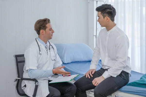 healthcare background of caucasian male medical doctor examining asian male patient sitting on bed in hospital