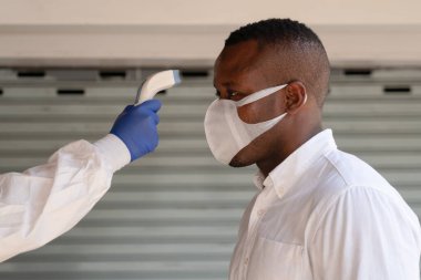 Koronavirüs covid-19 'daki ateşi kontrol etmek için Afrikalı Amerikalı iş adamlarına dijital cihazla virüs koruma giysileri ve yüz kalkanı tarama sıcaklığı