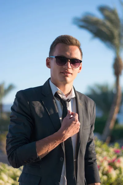 Young man stands in sunglasses and in a shirt at the palm tree — Stock Photo, Image