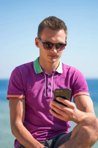 L'uomo in occhiali da sole si siede con il cellulare nelle mani vicino al mare — Foto Stock