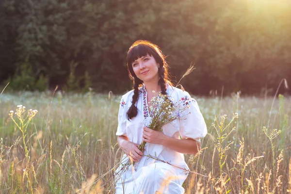 Молода жінка в довгій білій вишитій сорочці збирає квіти — стокове фото