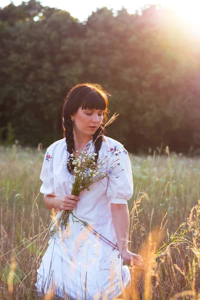 En ung kvinna i en lång vit broderad skjorta samlar blommor — Stockfoto