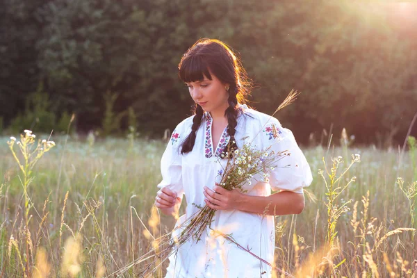 En ung kvinna i en lång vit broderad skjorta samlar blommor — Stockfoto