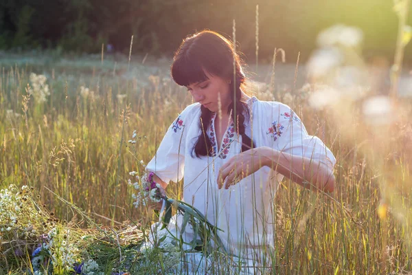 Молода жінка в довгій білій вишитій сорочці плете вінок — стокове фото