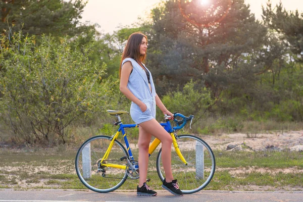 Mujer de pie con una bicicleta en una carretera en el parque al atardecer —  Fotos de Stock