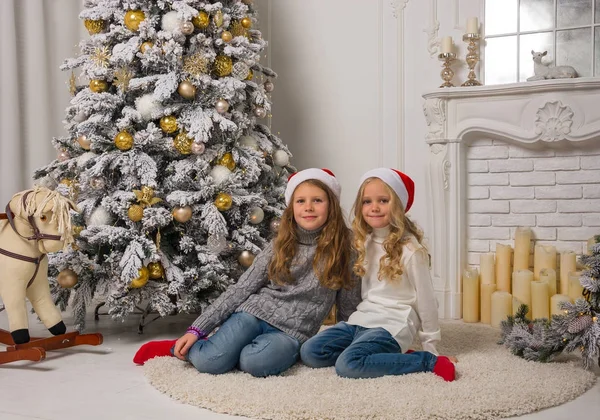 Two little pretty girls in red Christmas caps are sitting near t — Stock Photo, Image