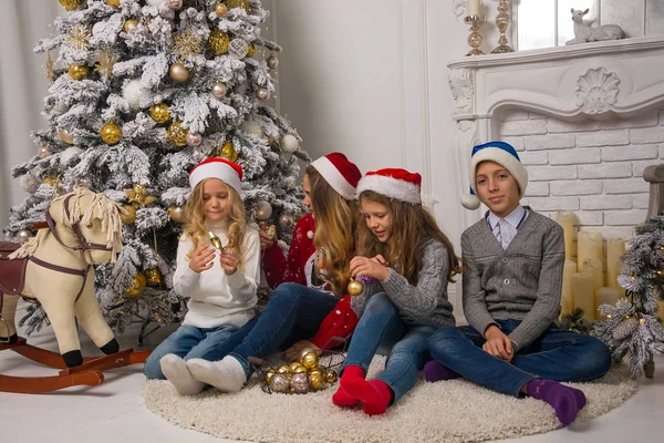 Kinder in Weihnachtsmützen schmücken einen Weihnachtsbaum in einem Raum — Stockfoto