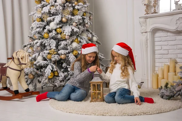 Dos niñas bonitas con gorras rojas de Navidad están sentadas cerca de t —  Fotos de Stock