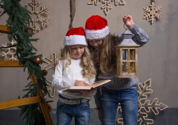 Two little girls in Santa caps are reading a book in a house — Stock Photo, Image