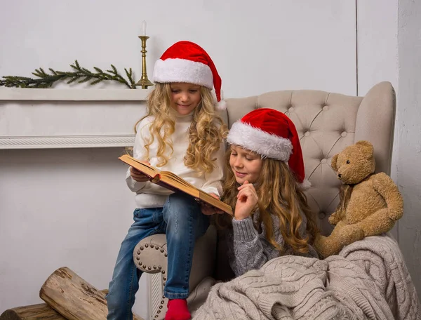 Two little girls in Santa caps are reading a book in the armchai — Stock Photo, Image