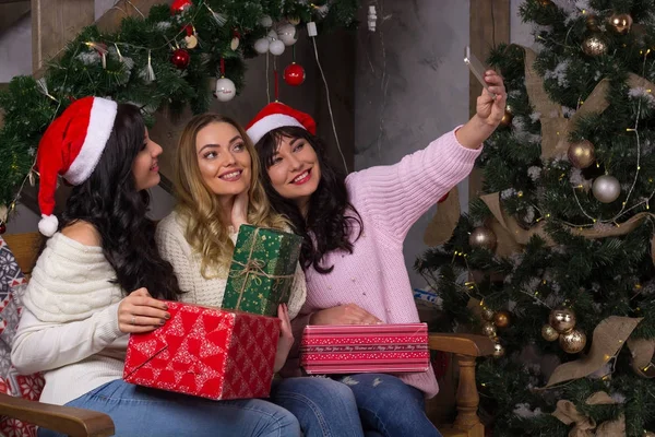 Tres hermosas mujeres en sombreros de santa sonrisas hacen selfies cerca de —  Fotos de Stock