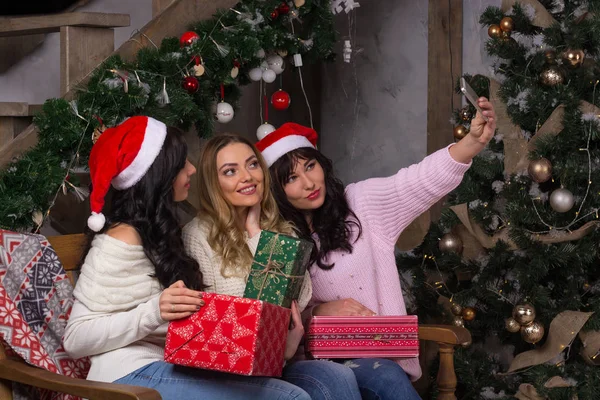 Three beautiful girls in santa hats smiles make selfies near of — Stock Photo, Image