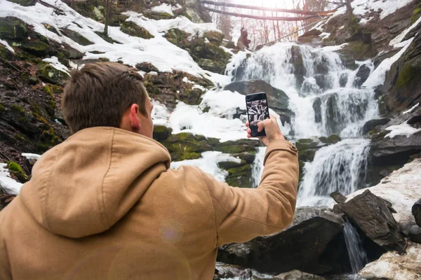 Um jovem tira uma foto de uma cachoeira em um telefone celular em — Fotografia de Stock