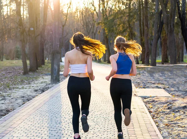 Young women run along the path in the park at sunset, back view