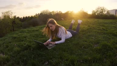 Genç güzel kız öğrenci bir dizüstü bilgisayar ve gülümsüyor çimlerin üzerine yaz aylarında günbatımı yatıyor.