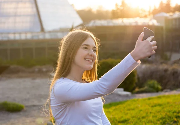 Uma jovem linda garota tirando selfie em um parque ao pôr do sol — Fotografia de Stock