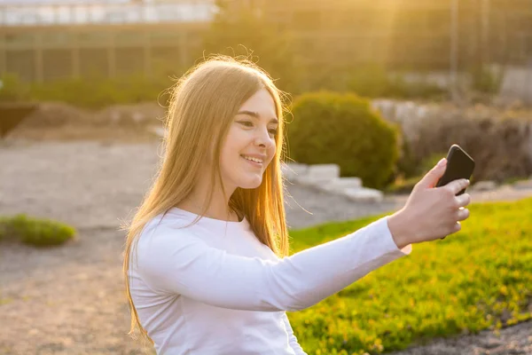 Uma jovem linda garota tirando selfie em um parque ao pôr do sol — Fotografia de Stock