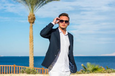 Takım elbiseli genç yakışıklı bir adam ve deniz dibinde beyaz pantolon ve palmiye ağaçları. Moda Modeli