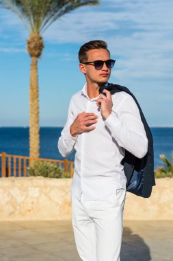 Takım elbiseli genç yakışıklı bir adam ve deniz dibinde beyaz pantolon ve palmiye ağaçları. Moda Modeli