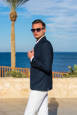 Takım elbiseli genç yakışıklı bir adam ve deniz dibinde beyaz pantolon ve palmiye ağaçları. Moda Modeli