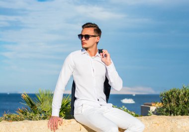 Takım elbiseli genç yakışıklı bir adam ve deniz dibinde beyaz pantolon ve palmiye ağaçları. Moda Modeli