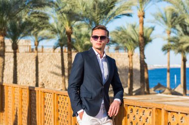 Beyaz gömlekli, beyaz pantolonlu, palmiye ağaçlarında ve plajda deniz altyapısı olan genç ve yakışıklı bir adam. Moda Modeli