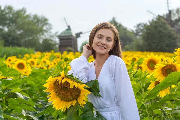 夏の田園風景を背景に ひまわり畑に白いシャツを着た美しい笑顔の若い女性が立っています — ストック写真