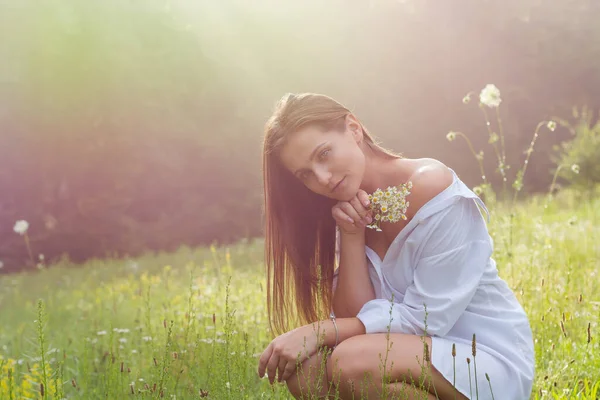 白いシャツを着た美しい若い女性が夏の森の風景を背景に牧草地で太陽の下で野生の花を開催しています — ストック写真