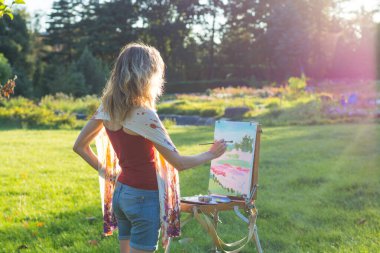Genç bir kadın sanatçı elinde bir fırça tutuyor ve gün batımının sehpasına resim çiziyor. Ressam gün batımında bahçede yağlı boya resimler yapar.