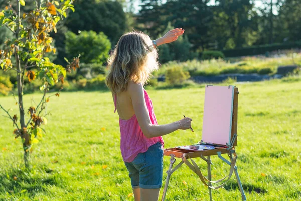 若い女性アーティストが筆を持ち 日没のイーゼルに絵を描く 画家は日没時に庭の油絵を描く — ストック写真