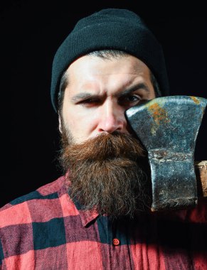 Şapkalı yakışıklı sakallı adam siyah arka planda balta tutuyor.
