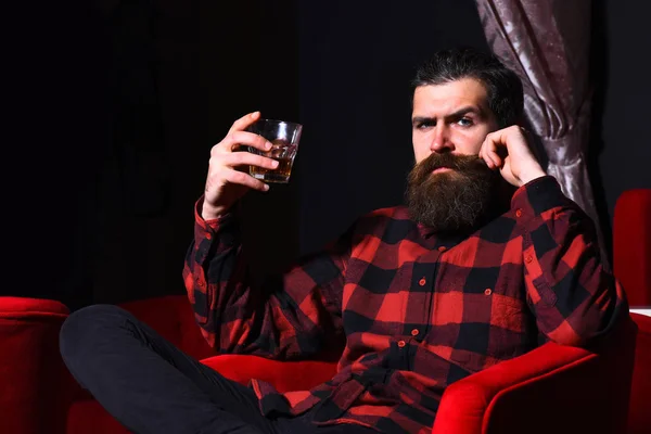 Brodaty człowiek hipster trzymając szklankę whisky w czerwonym krześle — Zdjęcie stockowe