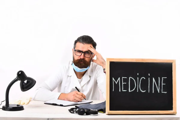 Médico caucasiano barbudo sentado à mesa com placa de inscrição "Medicina" — Fotografia de Stock