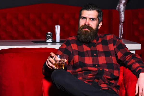 Brodaty człowiek hipster trzymając szklankę whisky w czerwonym krześle — Zdjęcie stockowe