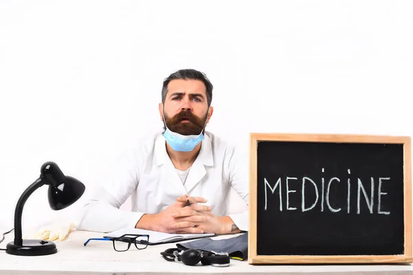 Skäggig kaukasisk läkare sitter vid bordet med ombord "Medicin" inskription — Stockfoto