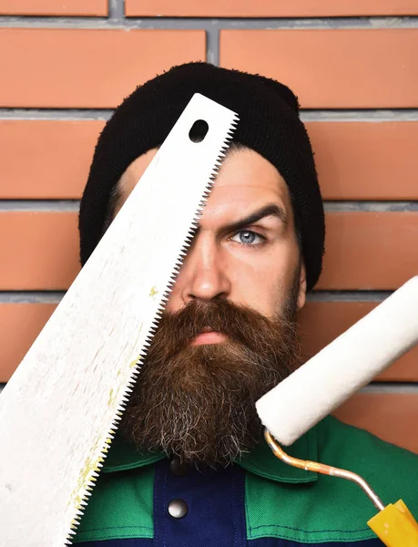 Бородатый рабочий держит различные строительные инструменты с серьезным лицом — стоковое фото