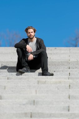 Mavi gökyüzü arkaplanına karşı basamaklarda oturan düşünceli genç adam
