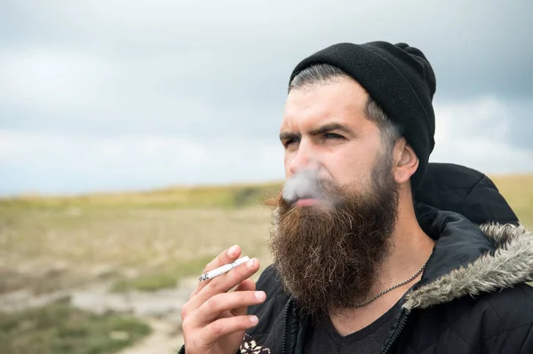 Bebaarde man rook sigaret in hoed buiten op natuurlijke achtergrond — Stockfoto