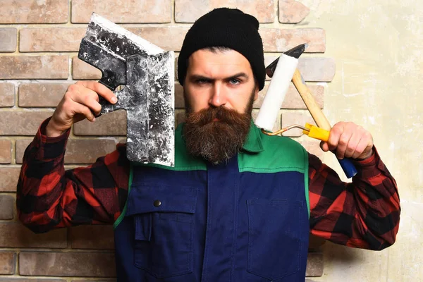 Homem construtor barbudo segurando várias ferramentas de construção com rosto sério — Fotografia de Stock