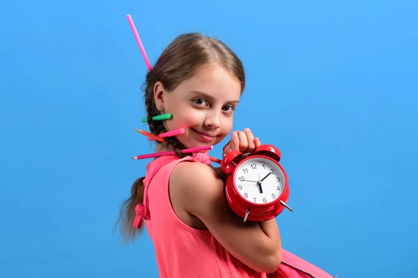 Kid in pink dress with colored pencils holds alarm clock — Stock Photo, Image