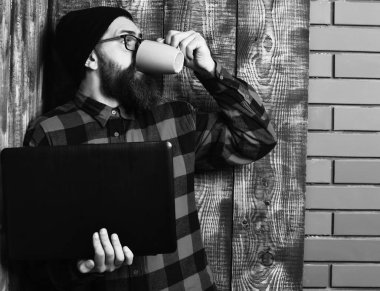 Sakallı, beyaz tenli hippi elinde bardakla dizüstü bilgisayar tutuyor.