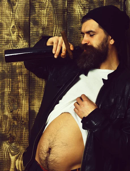 Бородатый брутальный кавказский хипстер держит бутылку и курит сигару. — стоковое фото