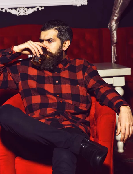 Γενειοφόρος hipster άνθρωπος κρατώντας ένα ποτήρι ουίσκι σε κόκκινη καρέκλα — Φωτογραφία Αρχείου