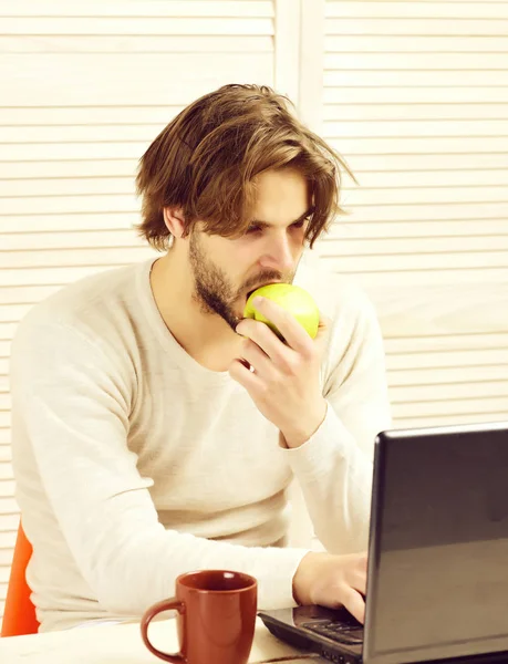 Guy uses his computer and eats apple. Man at work
