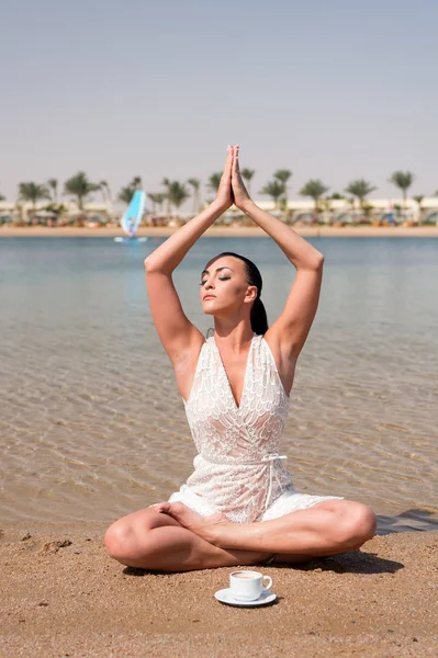Woman sitting in yoga pose with cup on sandy beach