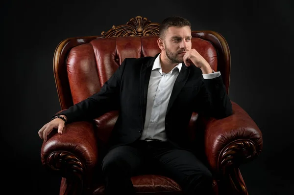 Man in formal outfit in leather chair on black background.