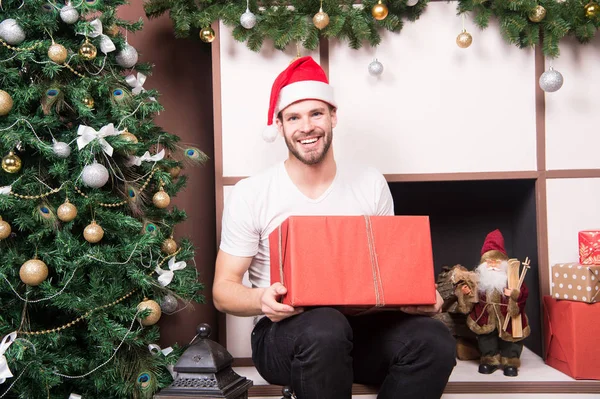Macho in red hat smile with wrapped box at fireplace — Stock Photo, Image